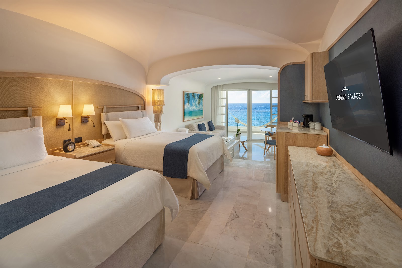 Spacious guest room at Cozumel Palace with two beds, ocean view balcony, flat-screen TV, and modern wood and marble furnishings.