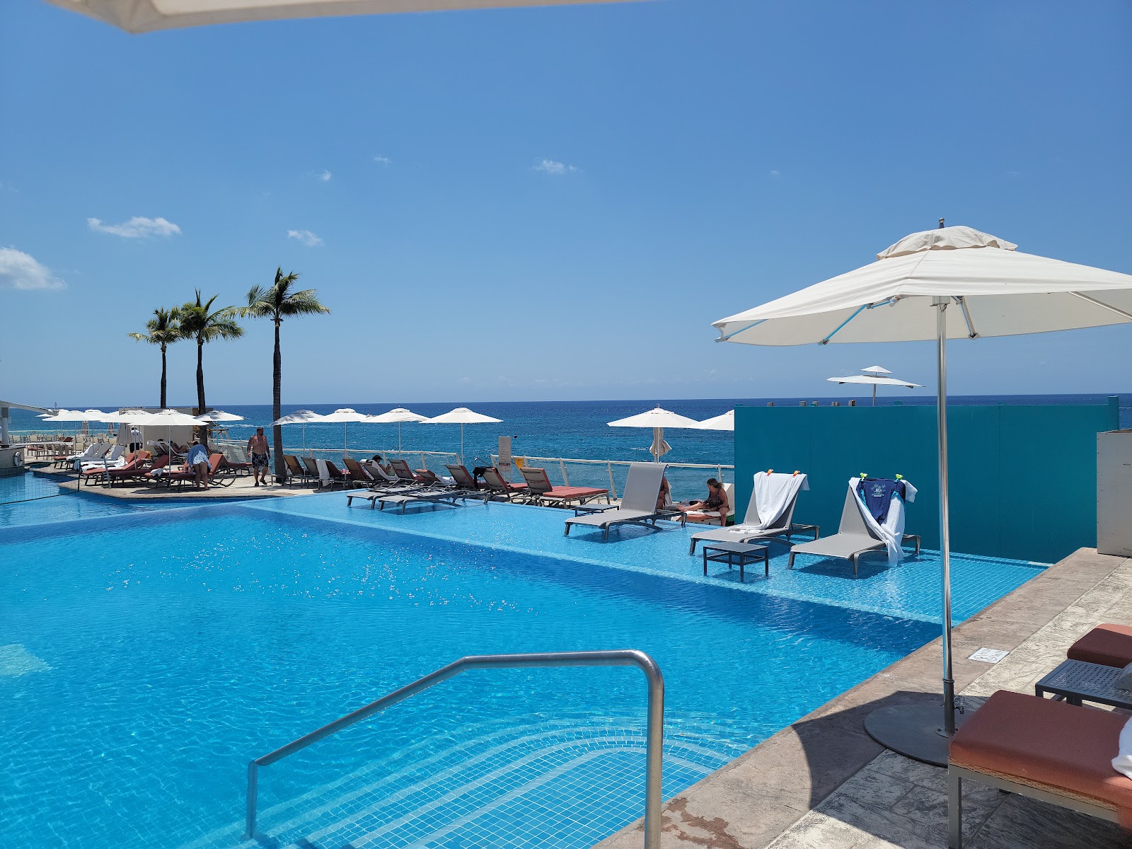 Large infinity pool with loungers and white umbrellas overlooking the Caribbean Sea at Cozumel Palace, palm trees visible beyond the pool deck.