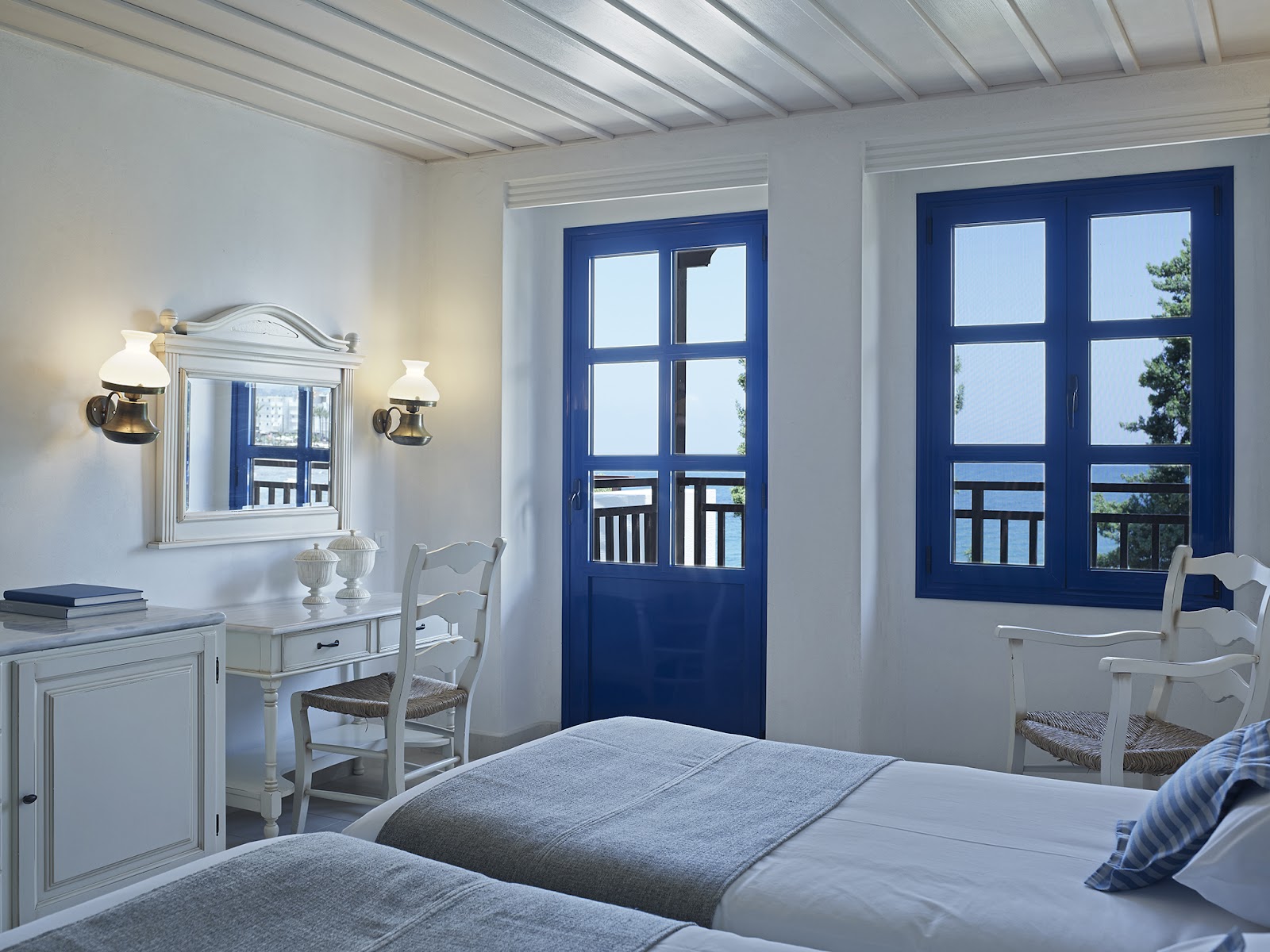 Bright resort bedroom with white bed, desk, and white chairs, opening to a balcony with sea view through blue-framed doors.