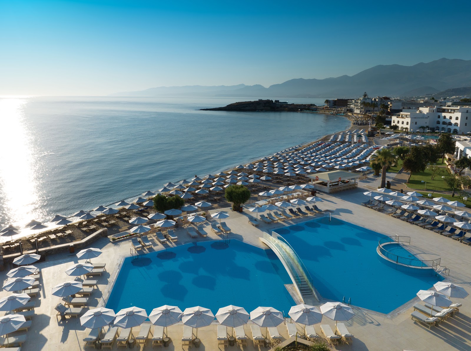 Resort pool with blue water and white umbrellas overlooking a Mediterranean beach and mountainous coastline.