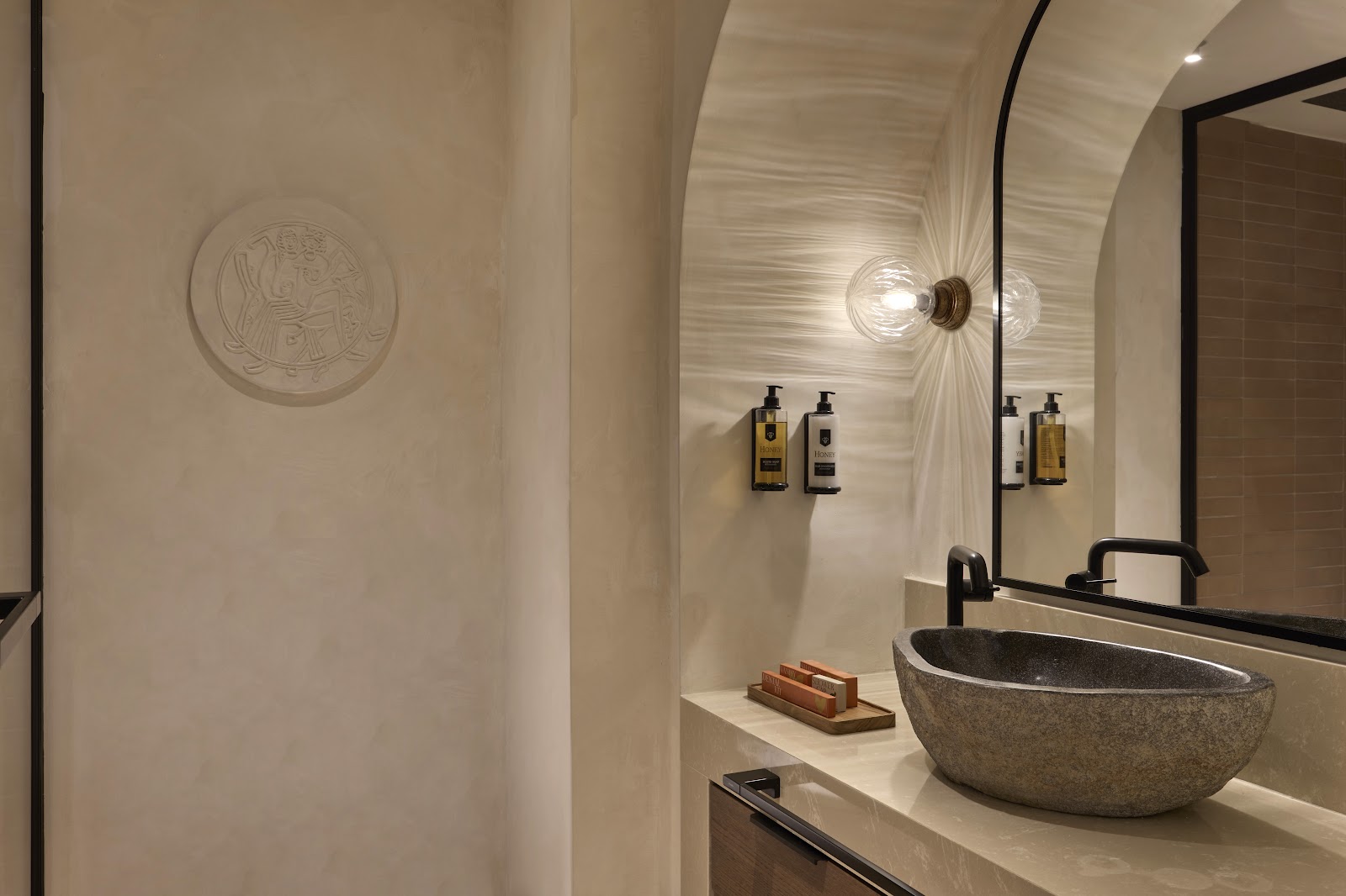 Modern bathroom with stone vessel sink, black-framed mirror, and amenity bottles under decorative wall sconce lighting.