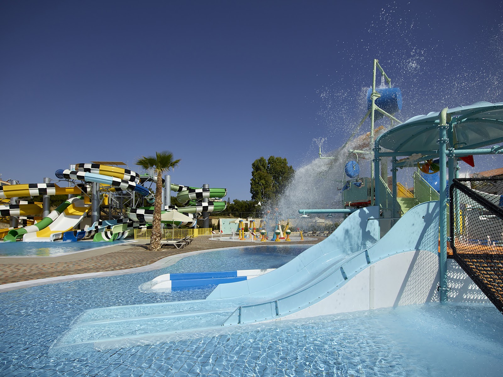Water slides and pools at Creta Maris Resort with checkered yellow and green slides, a bucket dumping water, and palm trees under clear blue sky.