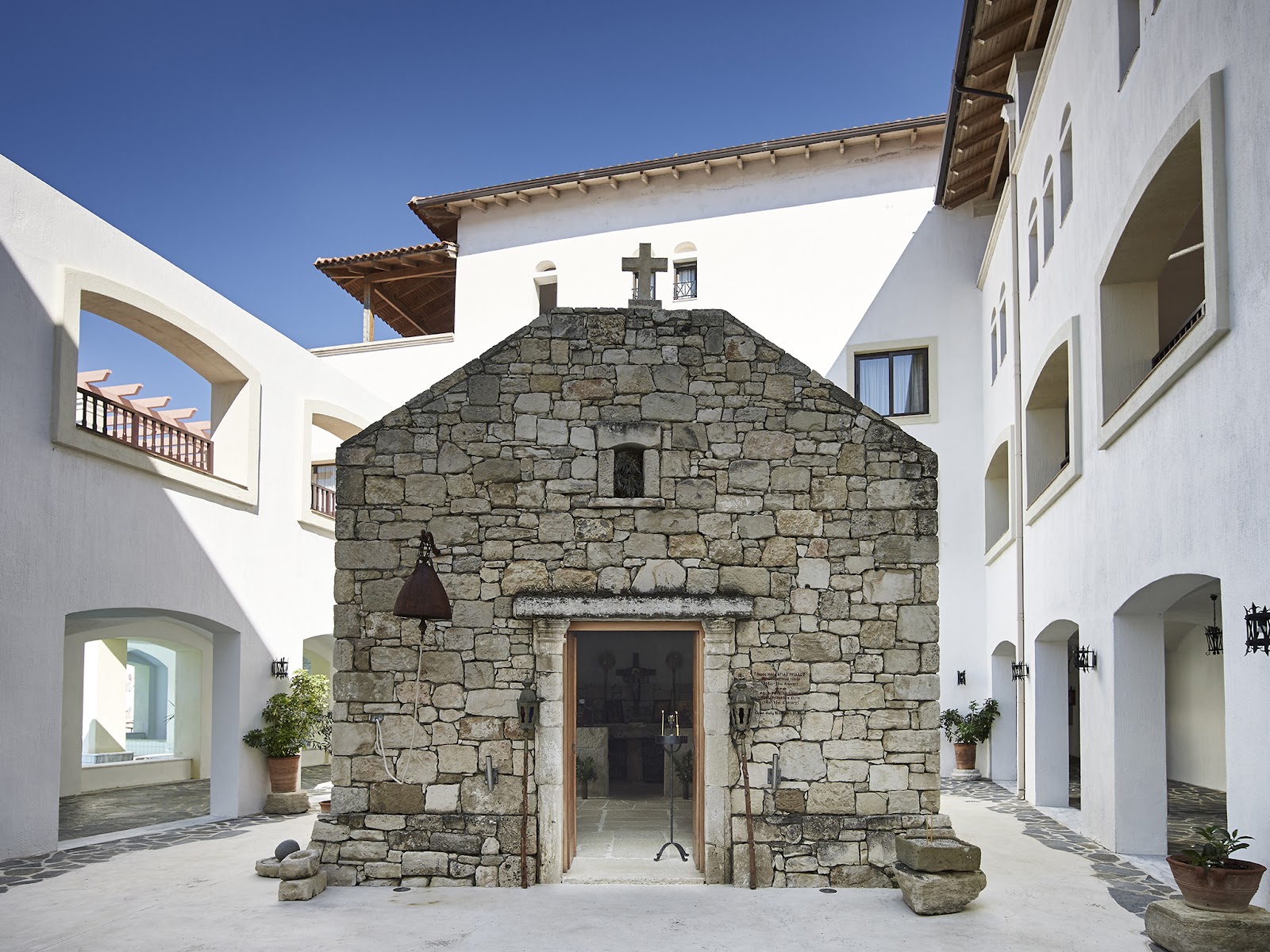 Historic stone chapel with cross at center of white courtyard surrounded by modern resort buildings under clear blue sky.