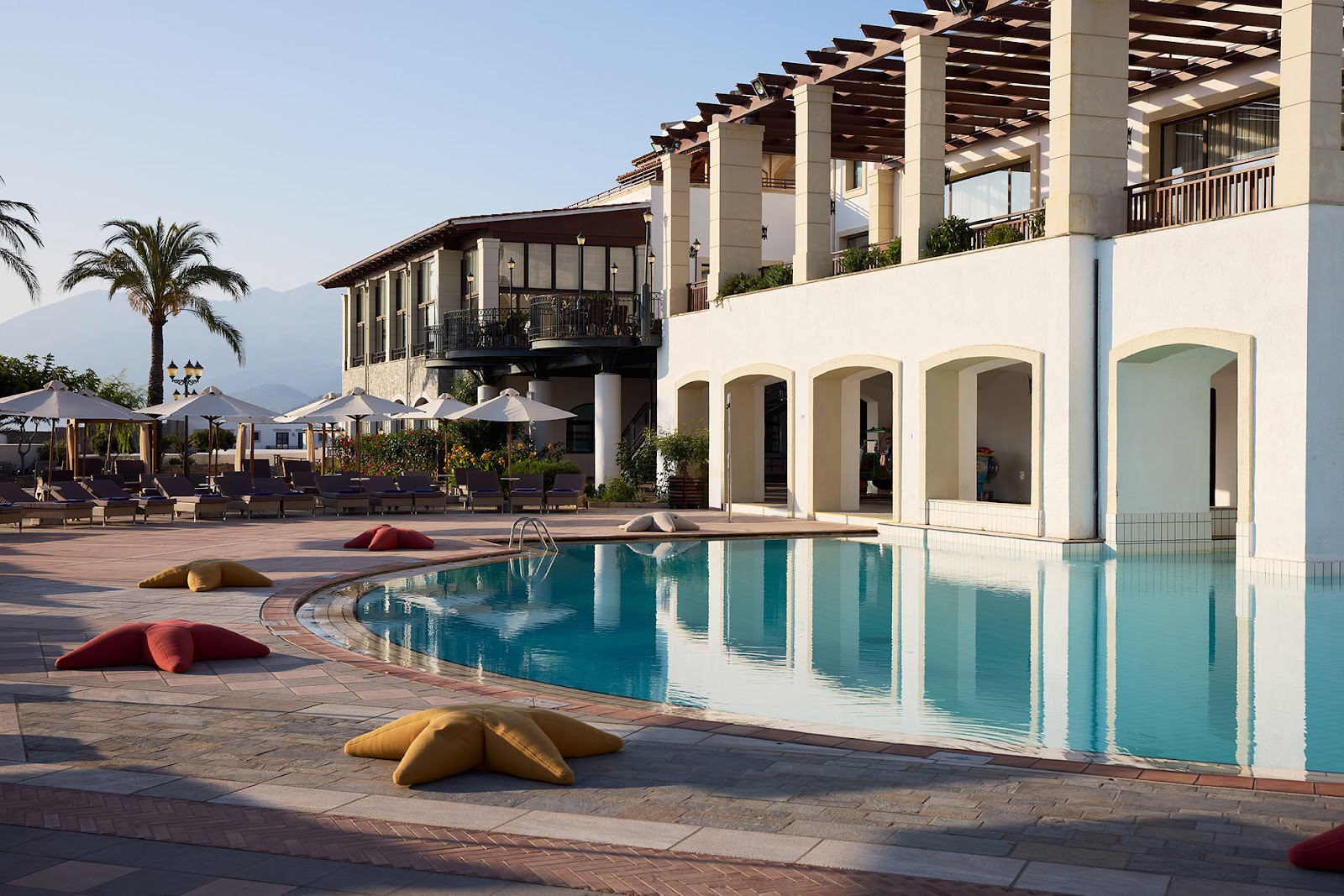 Spacious guest room with white furnishings, king bed with blue pillows, seating area, and French doors opening to a balcony overlooking the resort grounds.