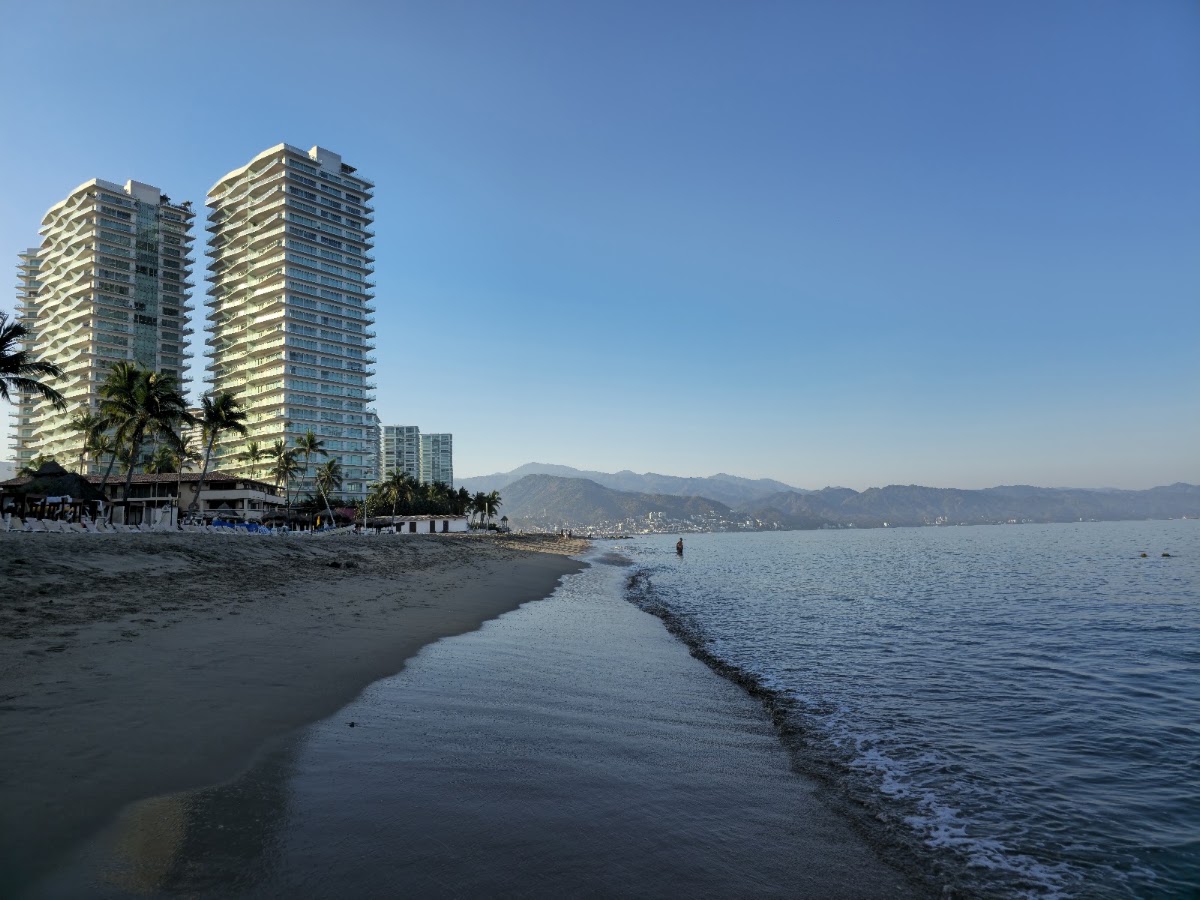 Sandy beach with gentle waves and two tall beige resort towers beside palm trees, mountains visible across the bay.