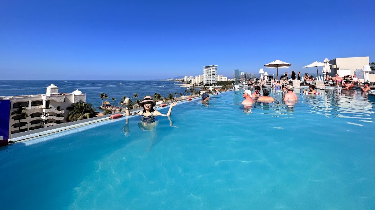 Guests swim in a bright turquoise rooftop pool overlooking the ocean and Puerto Vallarta coastline under clear blue sky.