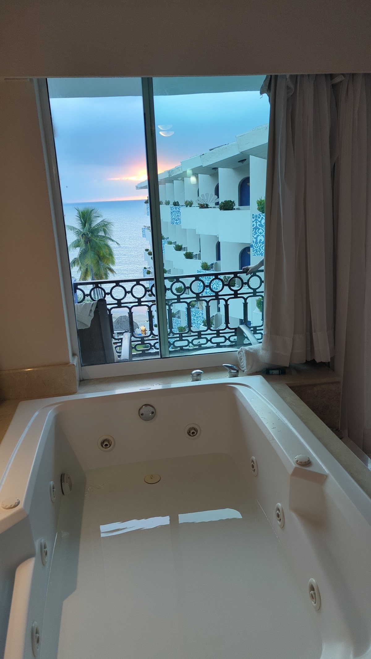 Jetted bathtub with ocean view from resort room balcony at sunset, white building and palm tree visible beyond.
