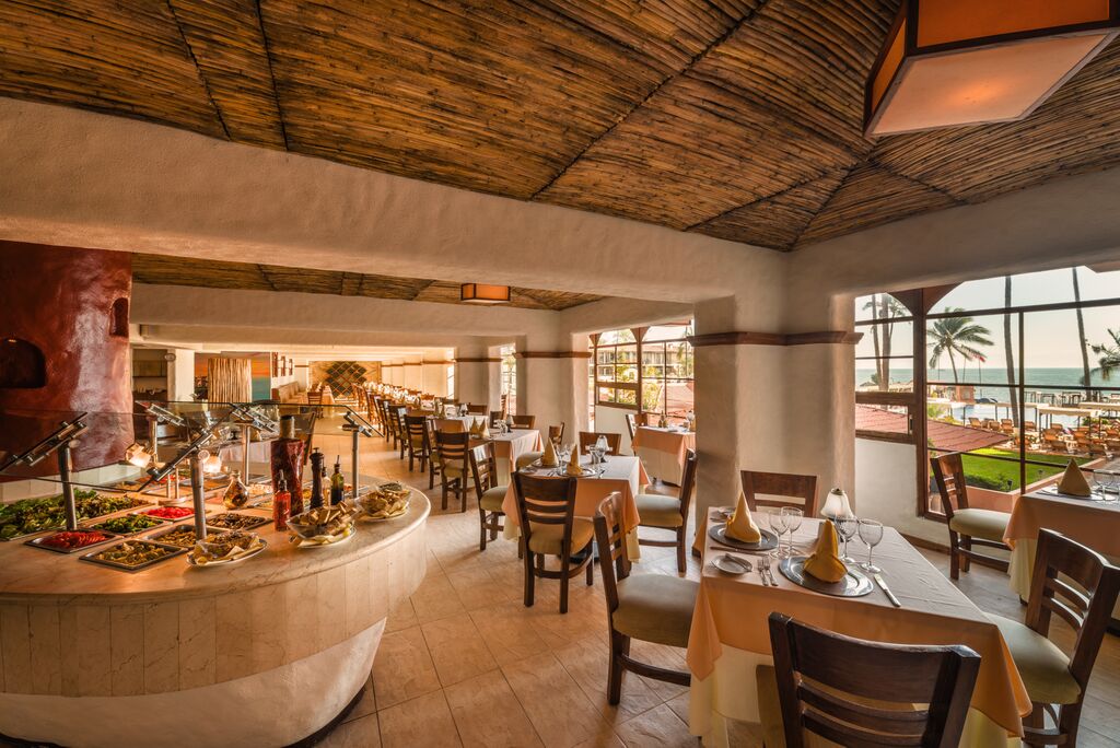Dining room with wooden ceiling, set tables, buffet counter with food displays, and ocean view through large windows.