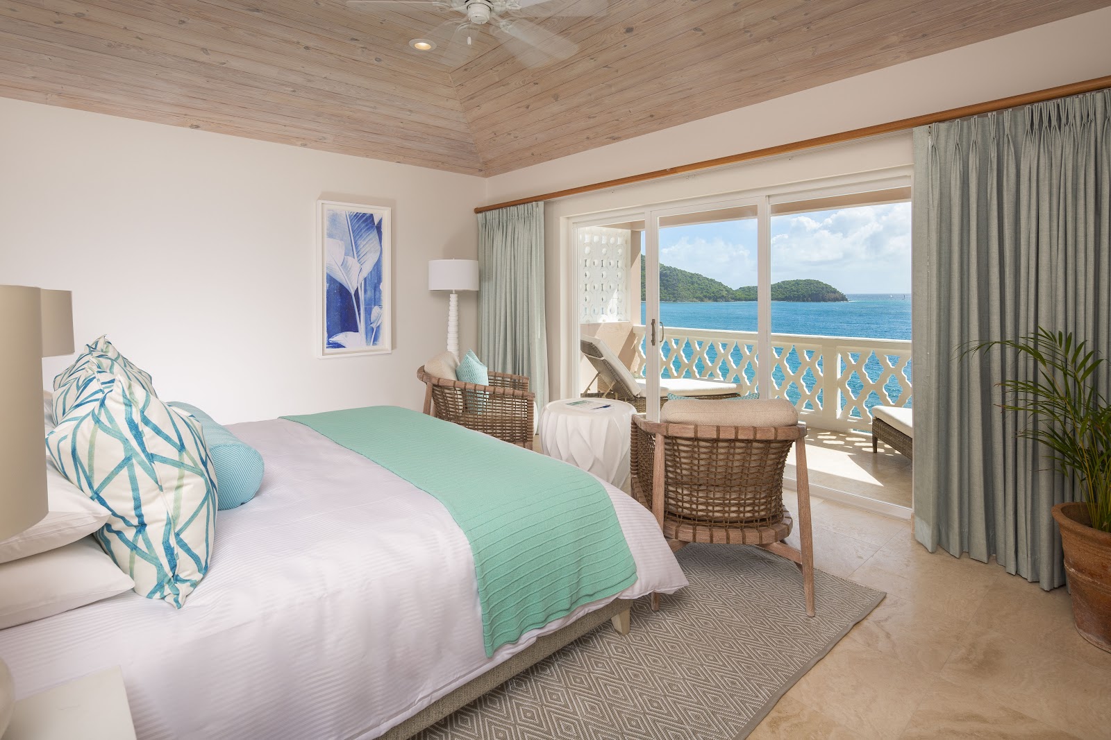Resort guestroom with ocean view balcony overlooking tropical islands and blue water, wood ceiling, queen bed with green throw, wicker seating, and glass doors.