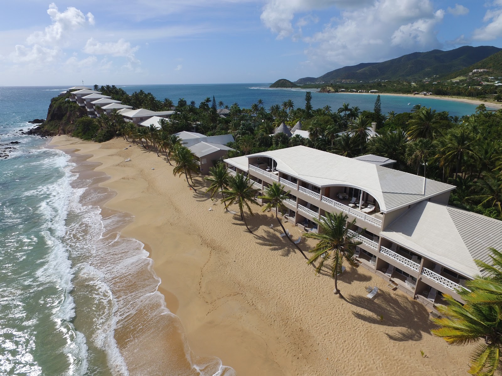 Modern white buildings with curved roofs sit on a sandy beach at Curtain Bluff, with palm trees, turquoise water, and a rocky headland visible along the Antigua coastline.