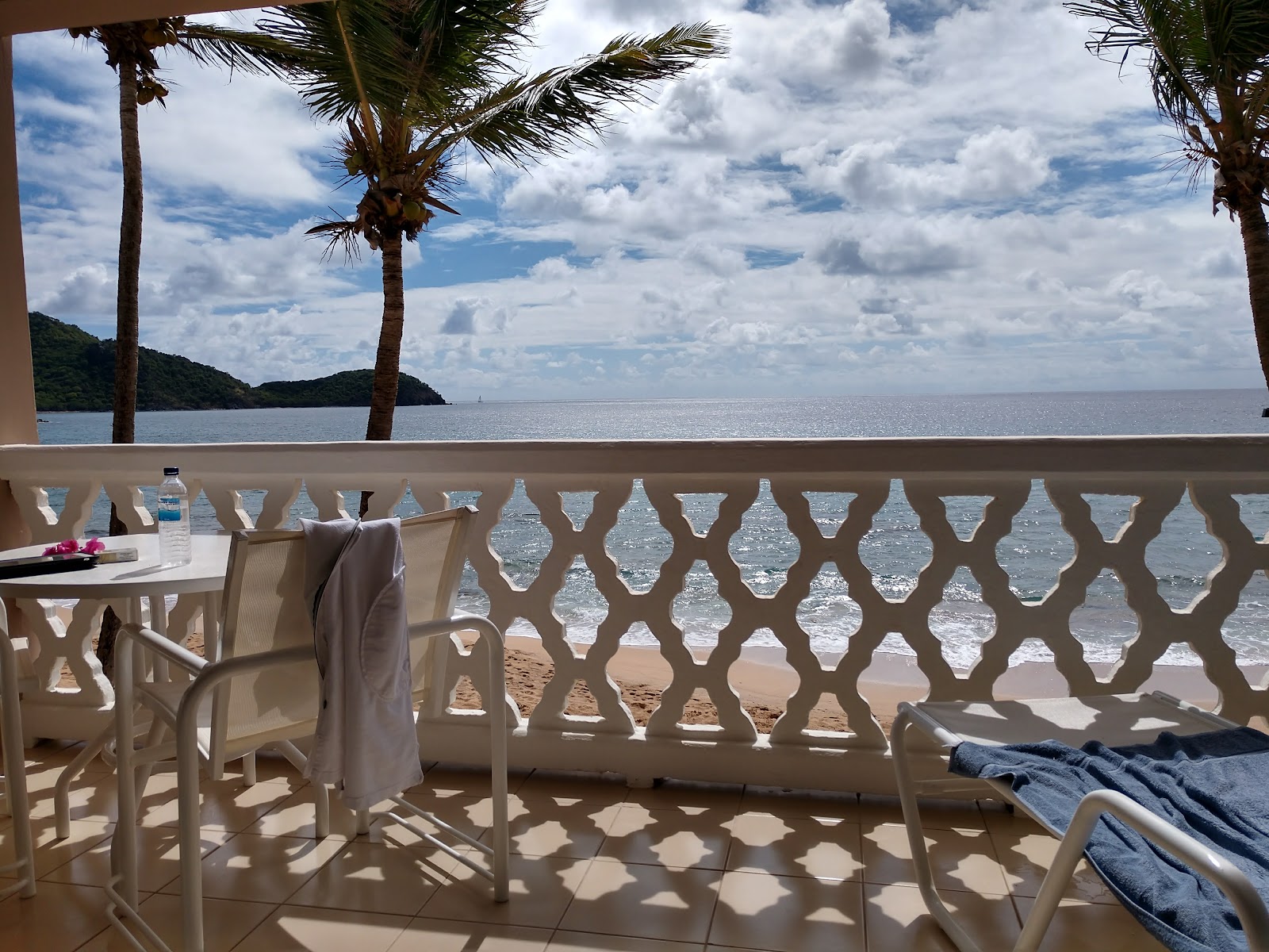 Beachfront balcony at Curtain Bluff with ocean view, palm trees, lounge chair, and dining table overlooking sandy beach and forested hillside.