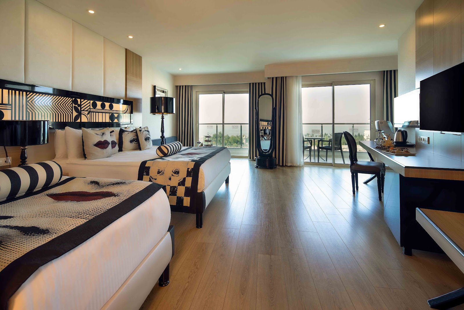 Spacious resort suite with two beds featuring black and gold patterned headboards, hardwood floors, and floor-to-ceiling windows overlooking landscaped grounds.