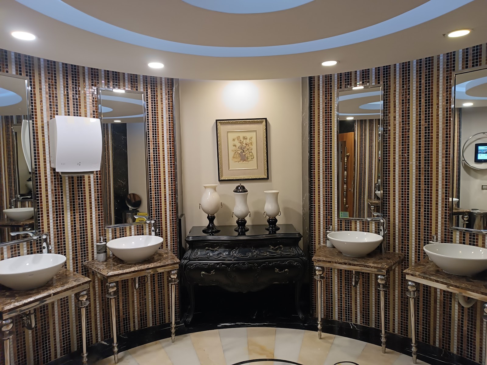 Spa bathroom with mosaic tile walls, multiple white vessel sinks, ornate black furniture, and framed botanical artwork at Delphin Imperial Hotel.