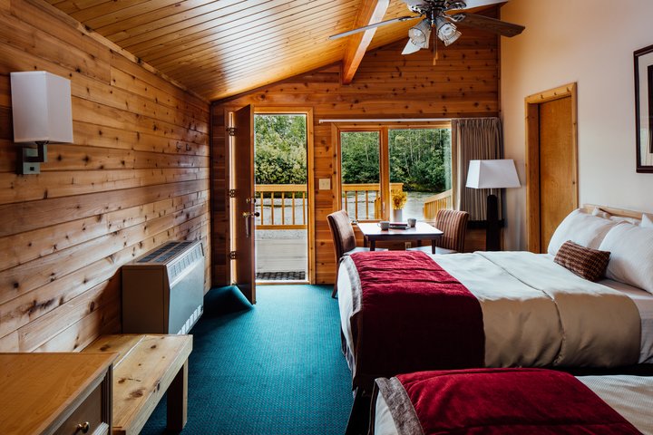 Spacious wooden lodge room with two beds, blue carpet, and glass doors opening onto a deck overlooking forested mountains.