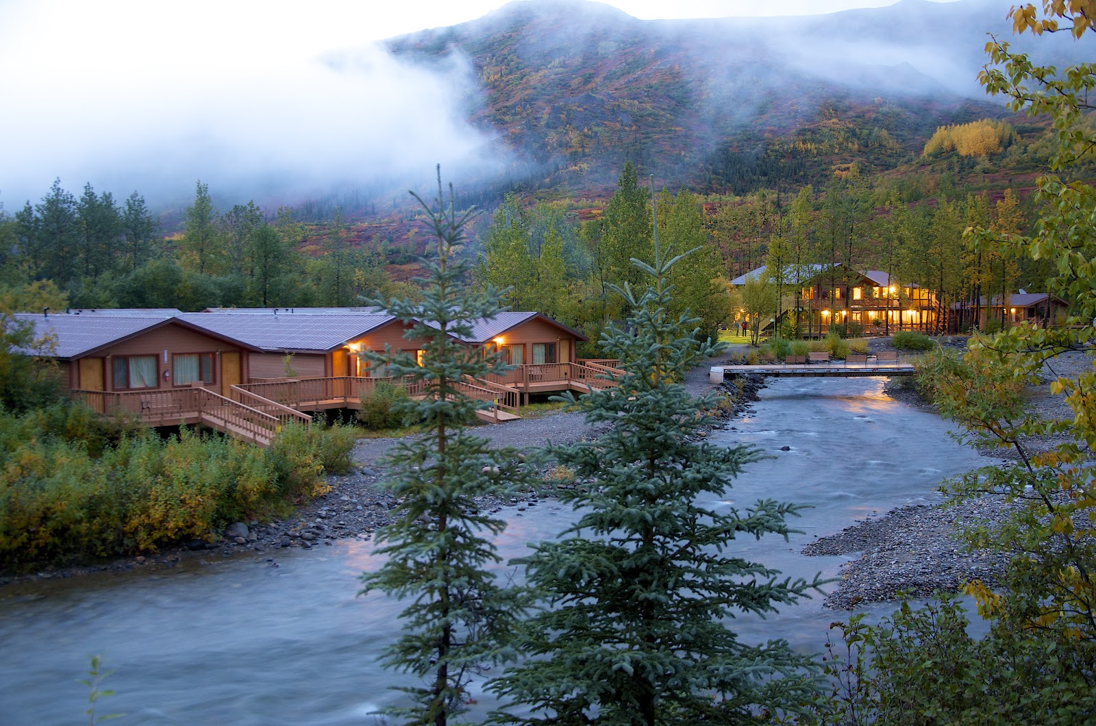 Denali Backcountry Lodge cabins with lit windows sit along a river at dusk, surrounded by coniferous forest and misty mountains.