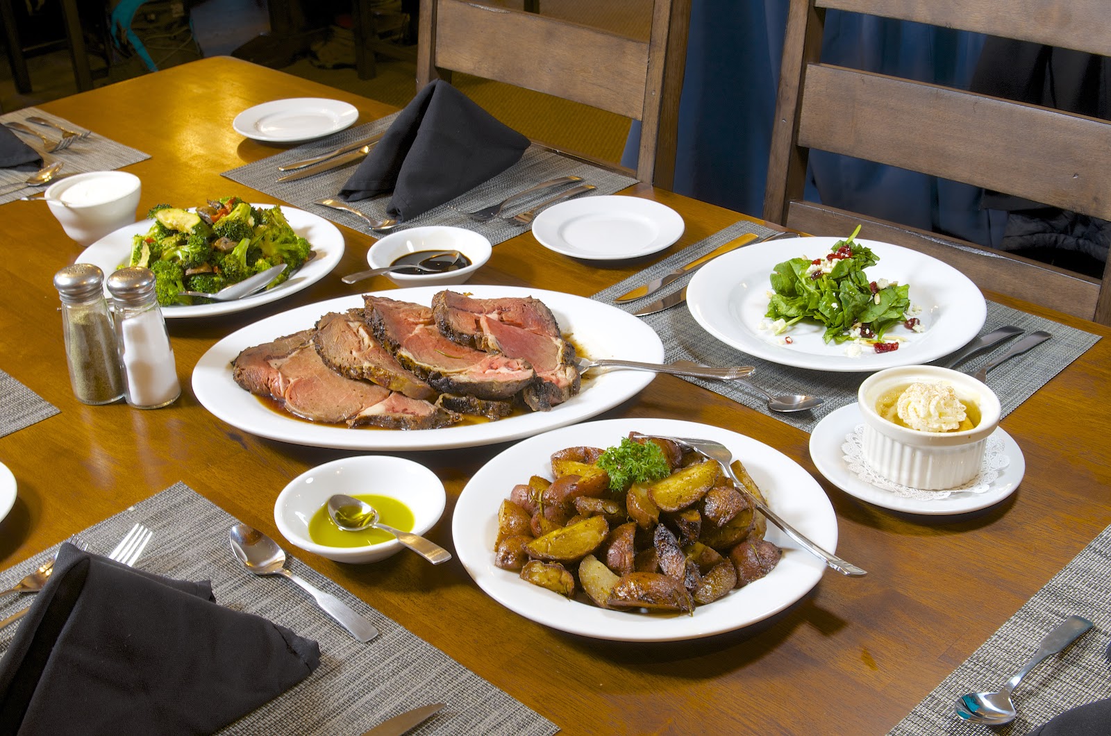 Spacious log cabin dining hall with wooden beams, antler chandelier, dark red chairs at round tables, and glass doors opening to wilderness views.