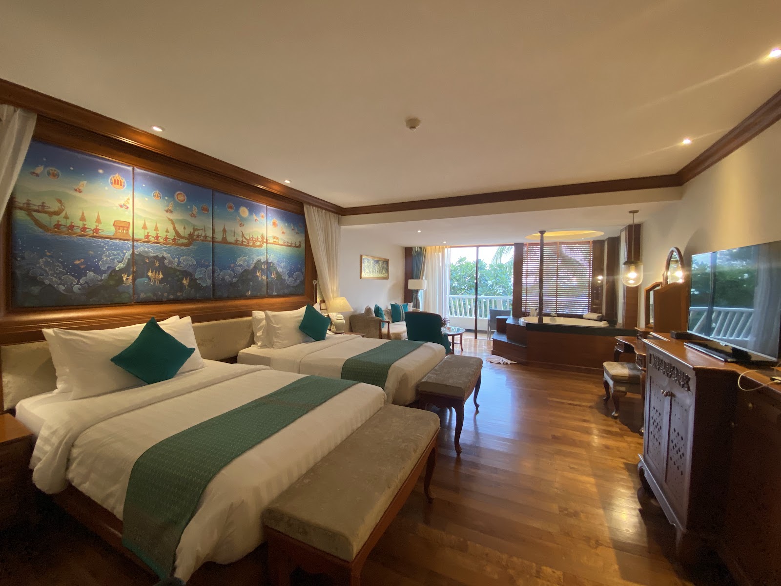 Spacious resort room with two queen beds, green runner blankets, and illuminated Thai temple artwork above the headboard, overlooking a balcony.