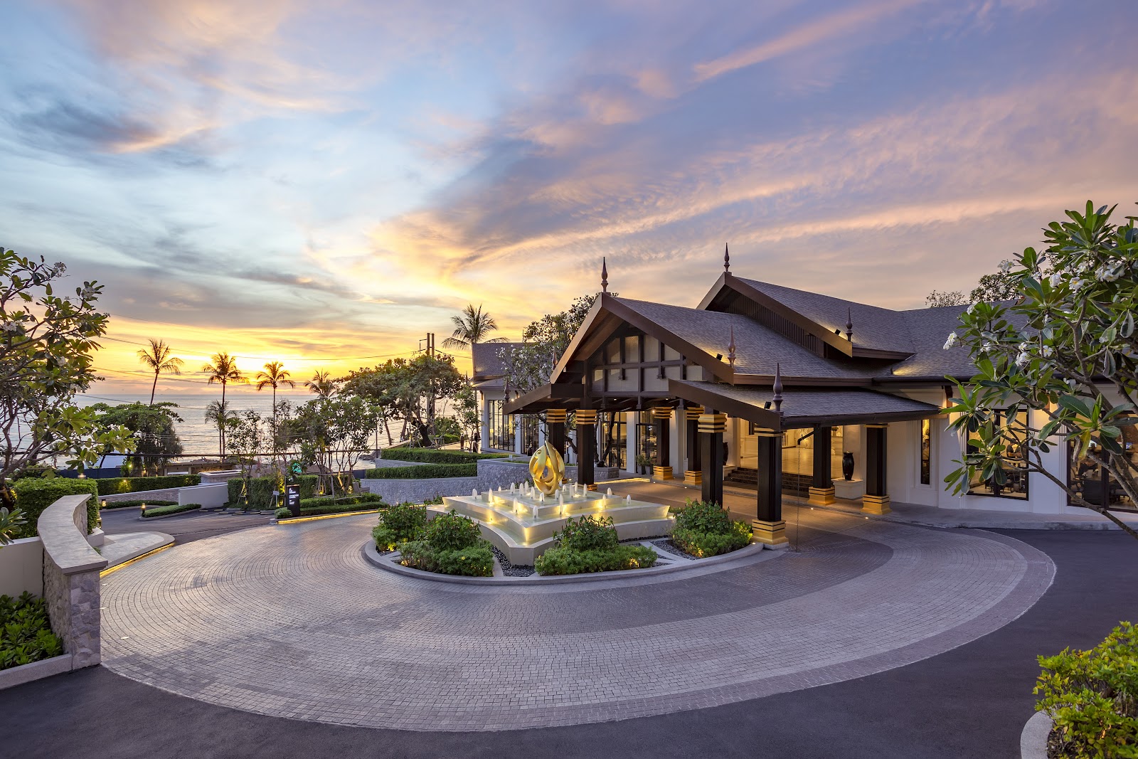 Diamond Cliff Resort & Spa's illuminated pavilion with peaked roof overlooks a courtyard fountain at sunset, palm trees and ocean visible beyond.