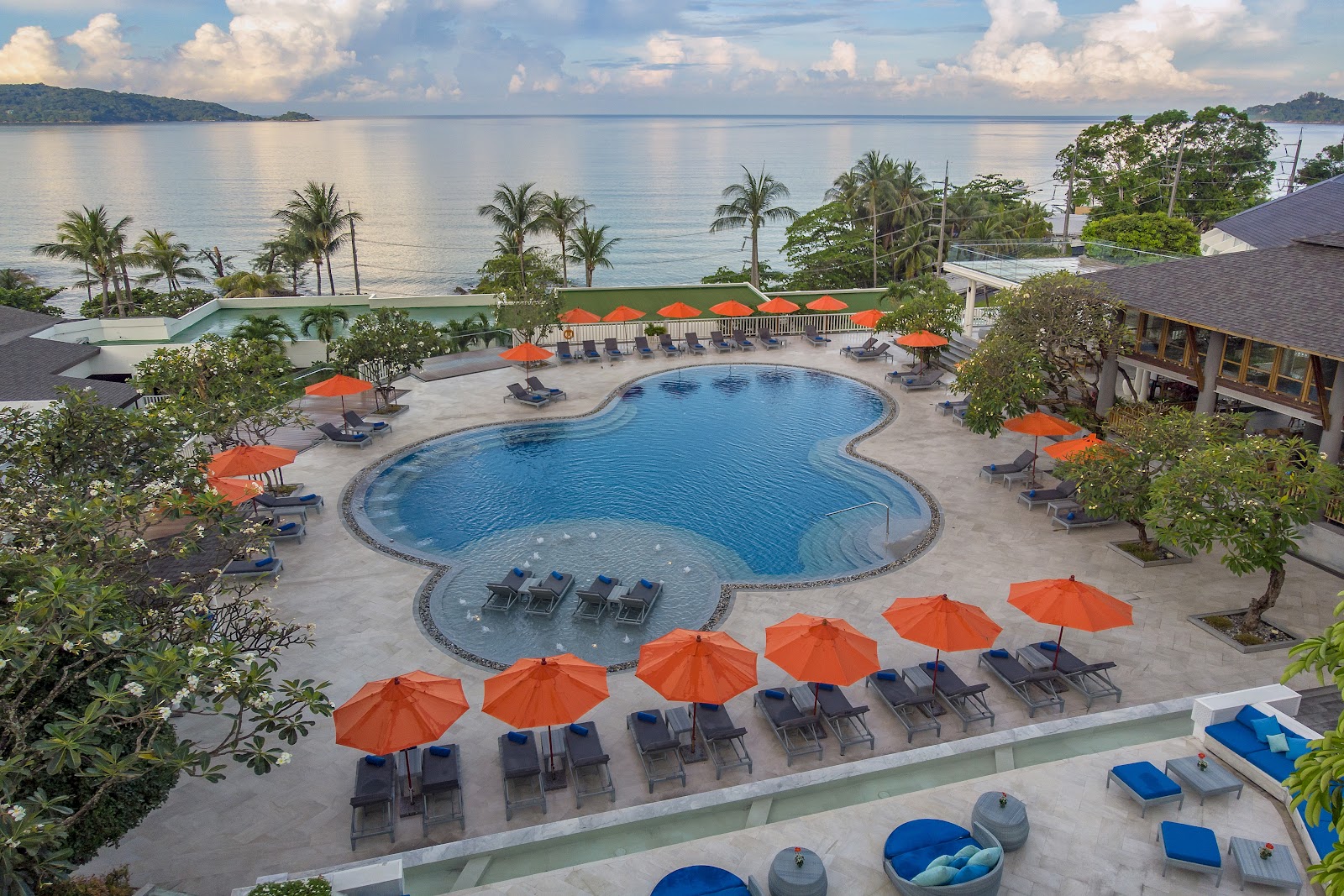 Resort pool with orange umbrellas and loungers overlooks calm bay with palm trees and forested hills under partly cloudy sky.