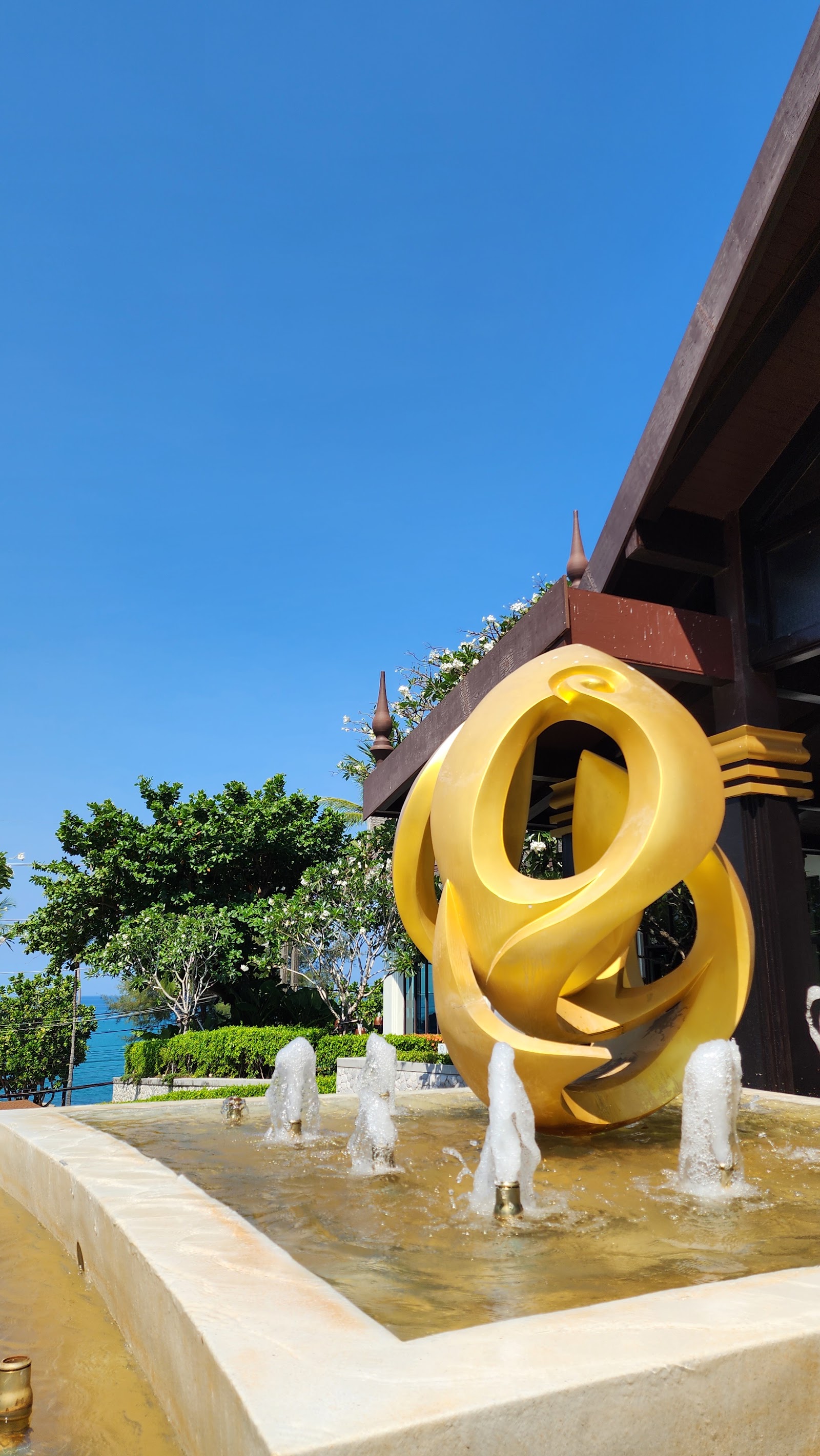 Large yellow sculptural installation in shallow water basin at Diamond Cliff Resort & Spa, with brown building and ocean view beyond under clear blue sky.