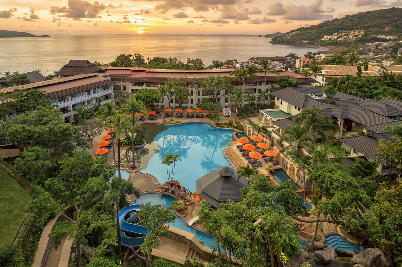 Diamond Cliff Resort & Spa overlooks a bay at sunset with its main pool, orange umbrellas, multi-story buildings, and water slide nestled among tropical vegetation.