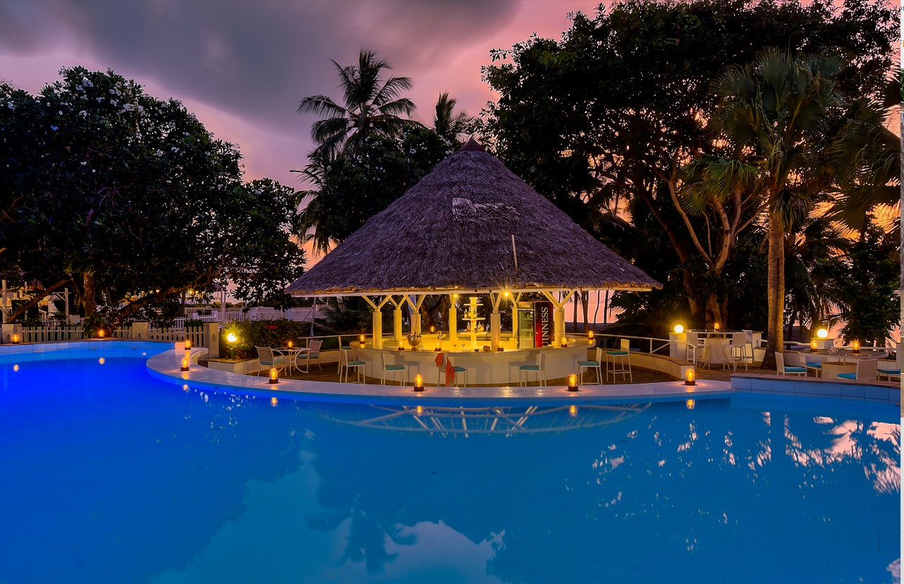 Illuminated thatched-roof bar pavilion beside a glowing blue pool at dusk, surrounded by palm trees and tropical vegetation.
