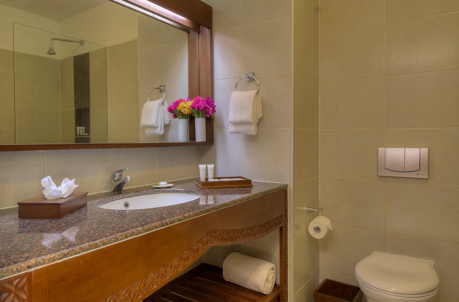 Resort bathroom with granite countertop, single sink, large mirror, white towels, pink flowers, and toilet with dark wood vanity.