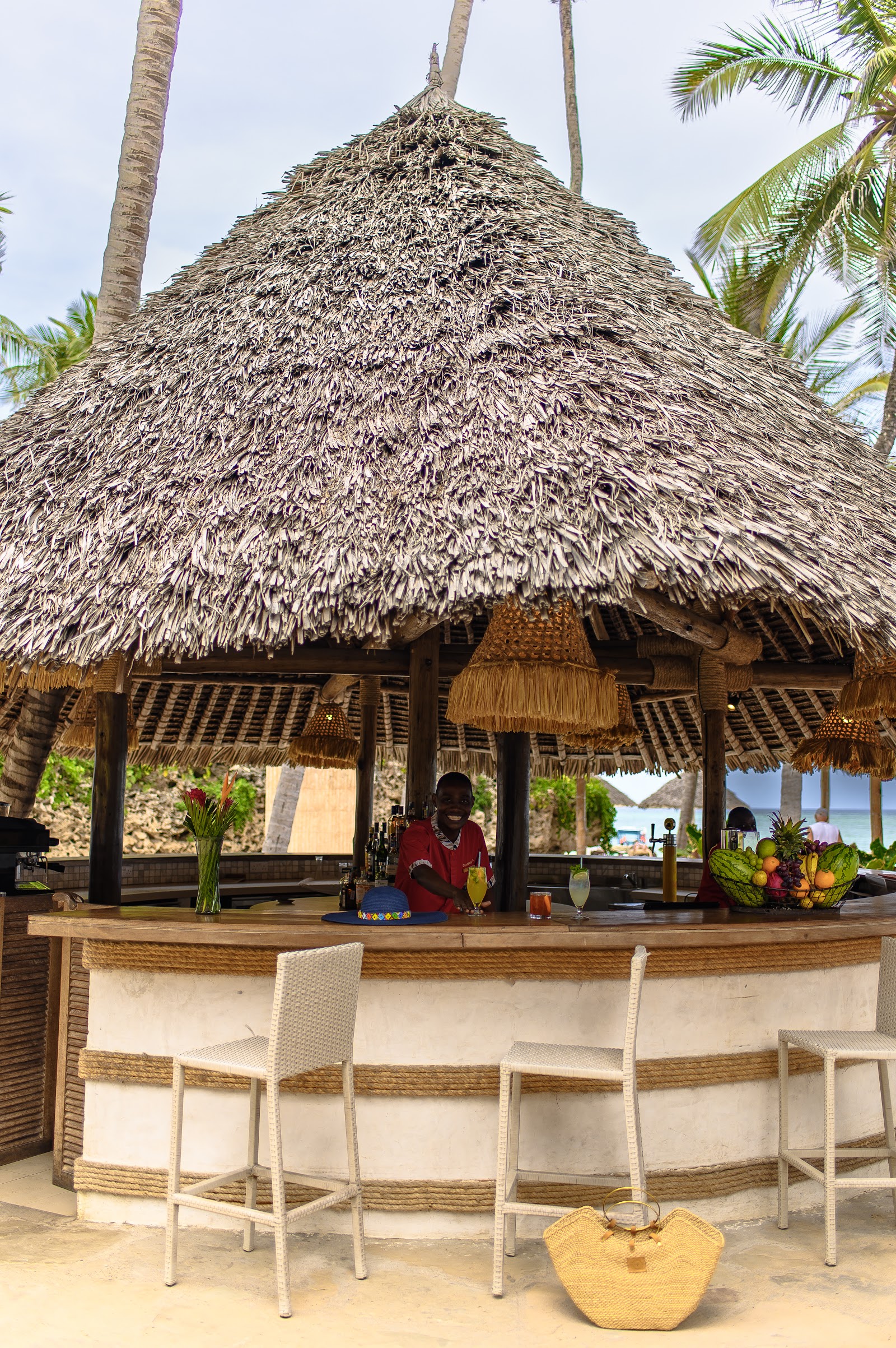 Thatched-roof beach bar at Diamonds Leisure Beach & Golf Resort with bartender serving drinks, woven chairs at counter, and ocean view beyond palm trees.