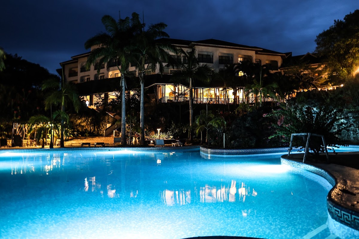 Illuminated pool complex at dusk with multi-level resort building, palm trees, and lounge chairs surrounding the glowing blue water.