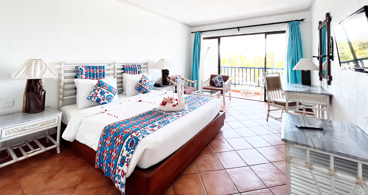 Spacious resort bedroom with king bed, turquoise curtains, ocean view balcony, and lounge seating area.