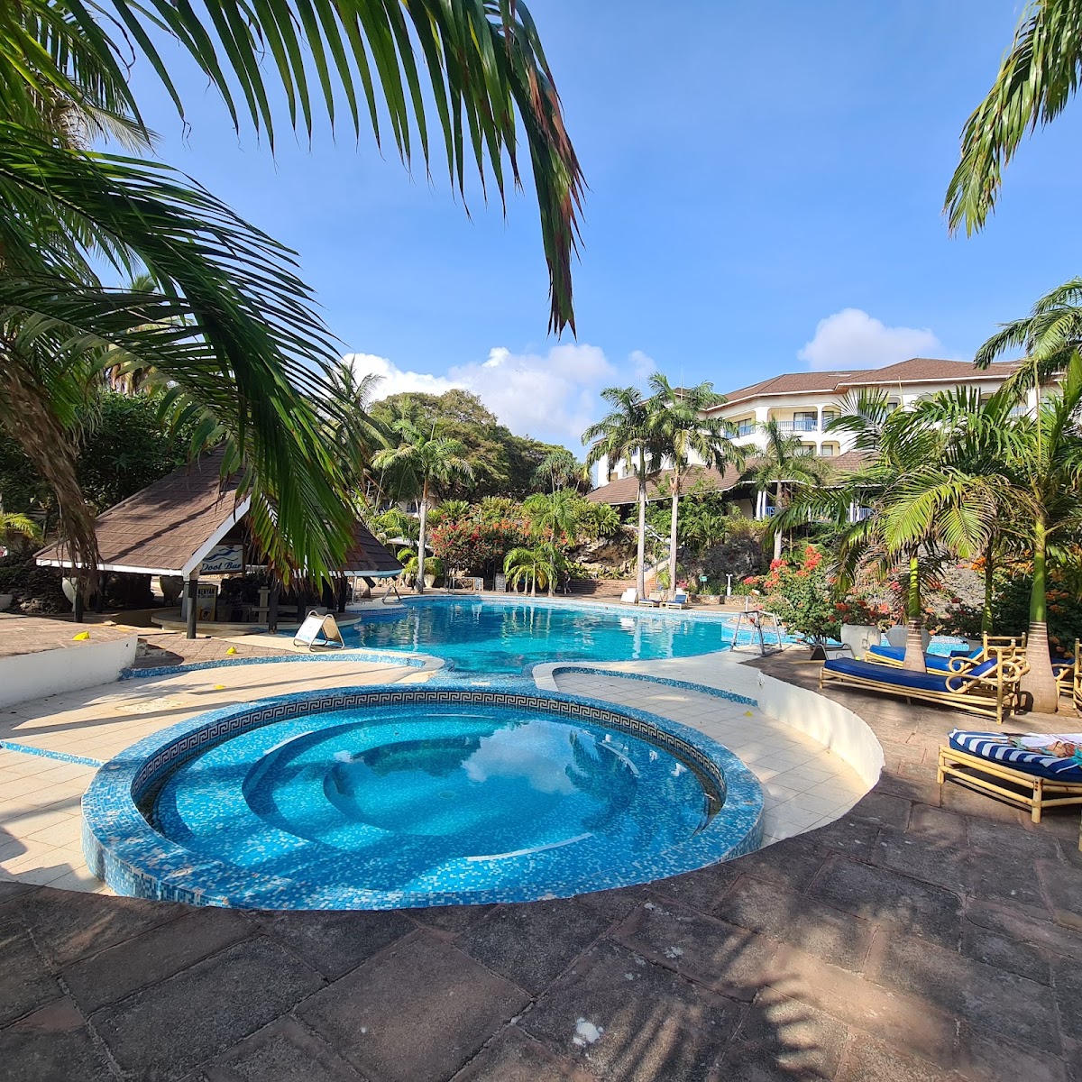 Circular hot tub in foreground with main swimming pool beyond, surrounded by palm trees, loungers, and a multi-story resort building under blue sky.