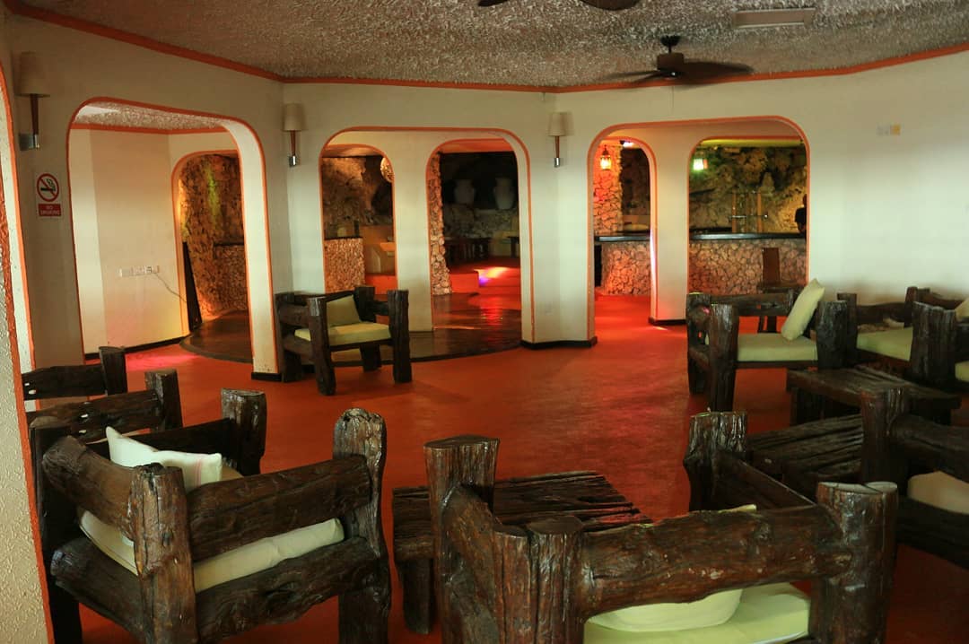 Lounge area with red-orange flooring, rustic wooden chairs with green cushions, and rounded doorways with warm lighting leading to other spaces.