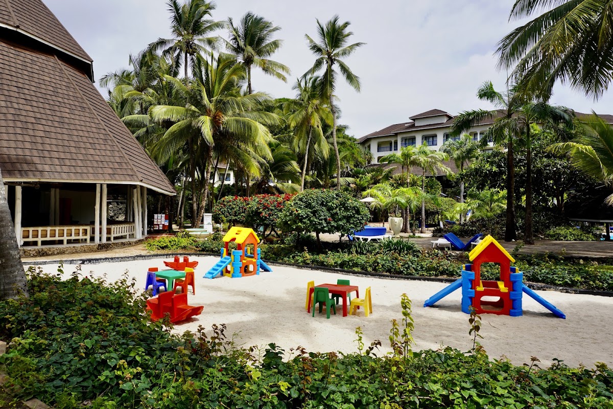 Children's play area with colorful playground structures on sand, surrounded by palm trees and resort buildings at Diani Reef Beach Resort & Spa.