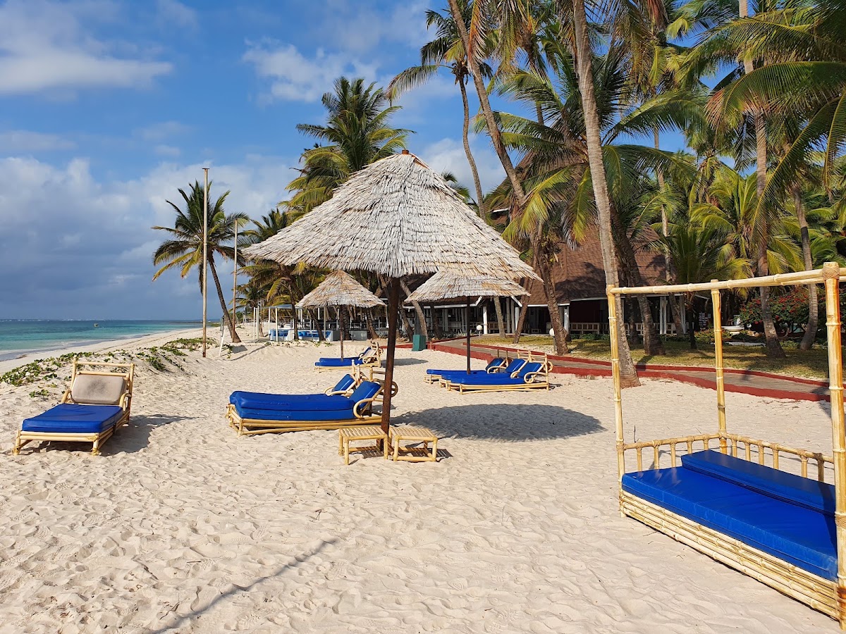 Blue loungers and thatched umbrellas dot the sandy beach at Diani Reef Beach Resort & Spa, with palm trees and turquoise ocean beyond.