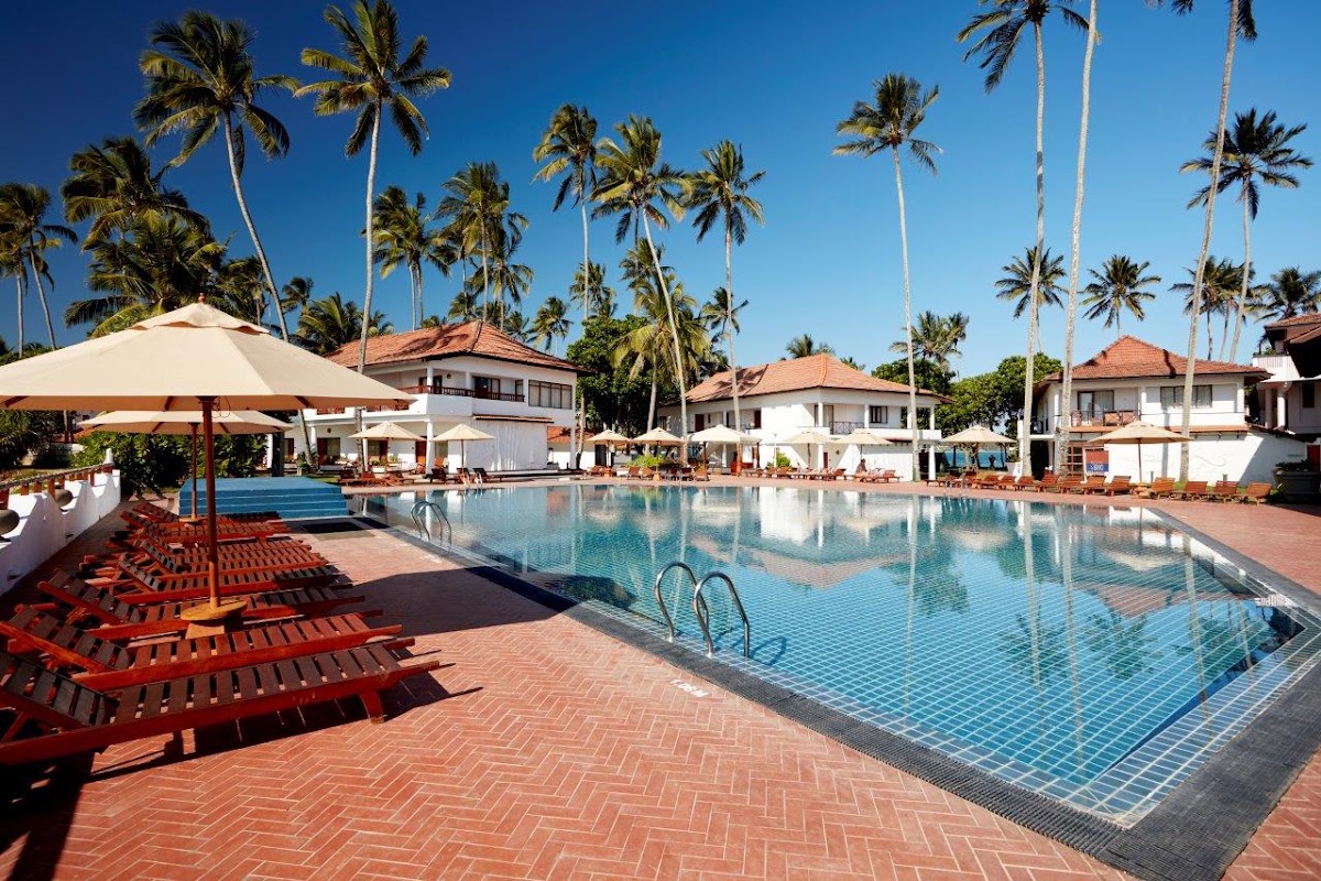 Red lounge chairs and umbrellas line the brick deck beside a large swimming pool at Dickwella Resort and Spa, with white bungalows and tall palms beyond.