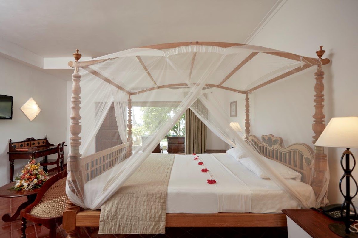 Four-poster canopy bed with white drapes and red rose petals at Dickwella Resort and Spa, with wooden furniture and garden view.