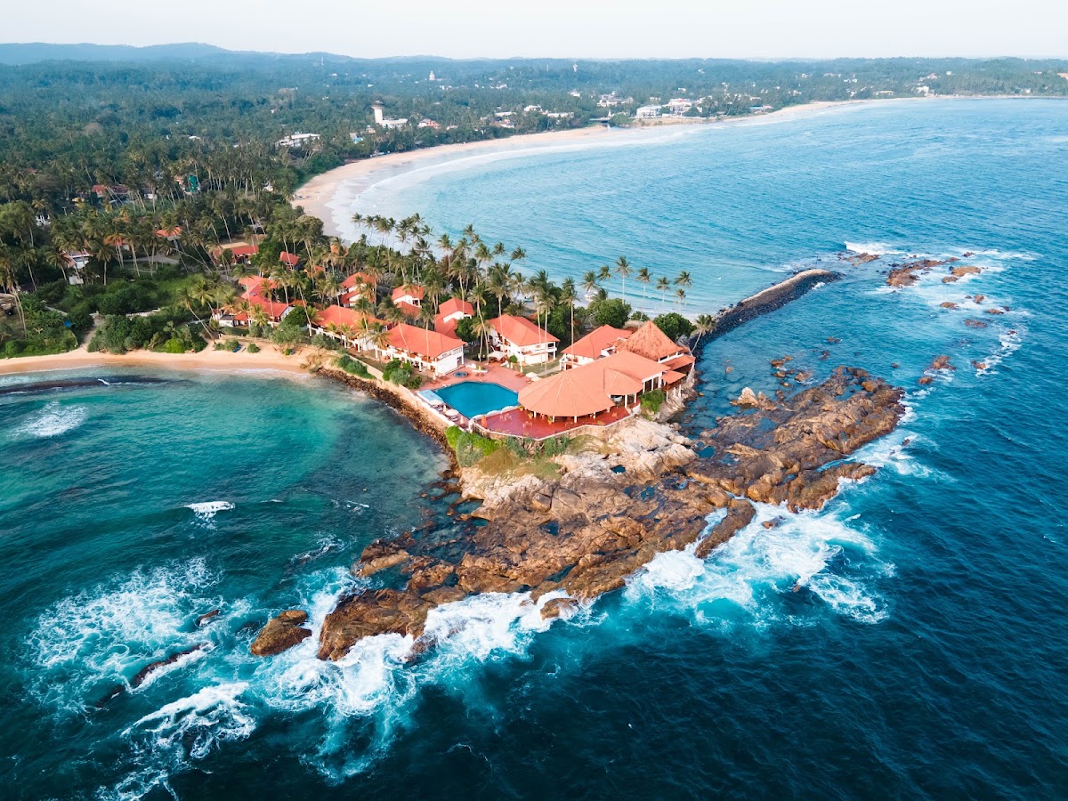 Aerial view of Dickwella Resort and Spa on a rocky peninsula with red-roofed buildings, a pool, and a sandy beach backed by forested hills.