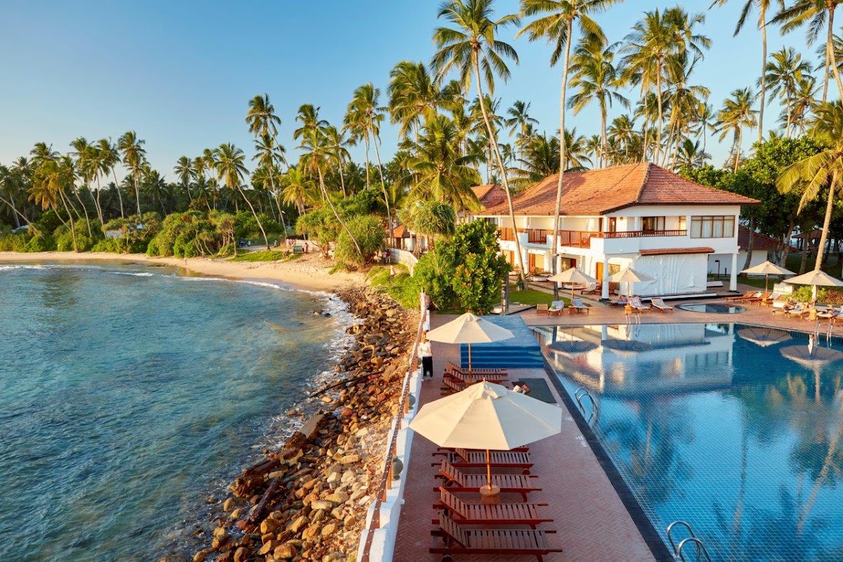 Dickwella Resort and Spa sits on a rocky coastline with a pool, umbrellas, and lounge chairs overlooking turquoise water and a sandy beach lined with palm trees.