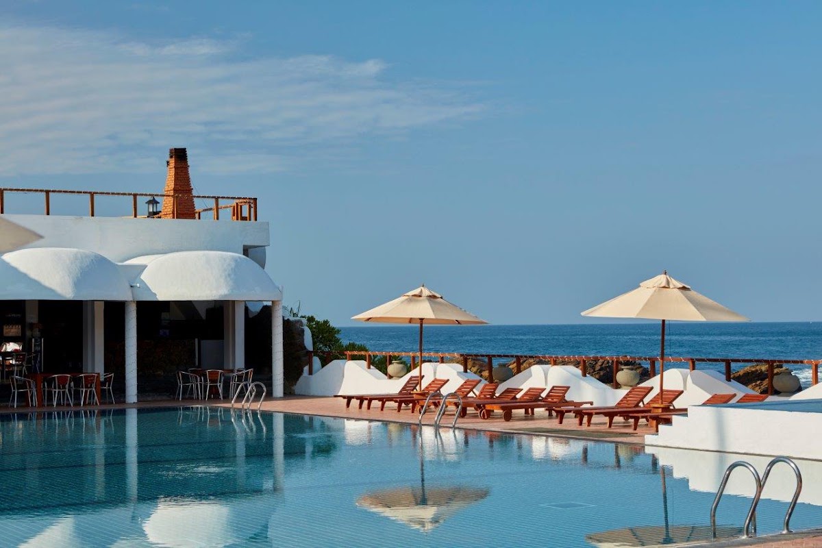 Pool area at Dickwella Resort and Spa with loungers, umbrellas, bar pavilion, and ocean view beyond rocky coastline.