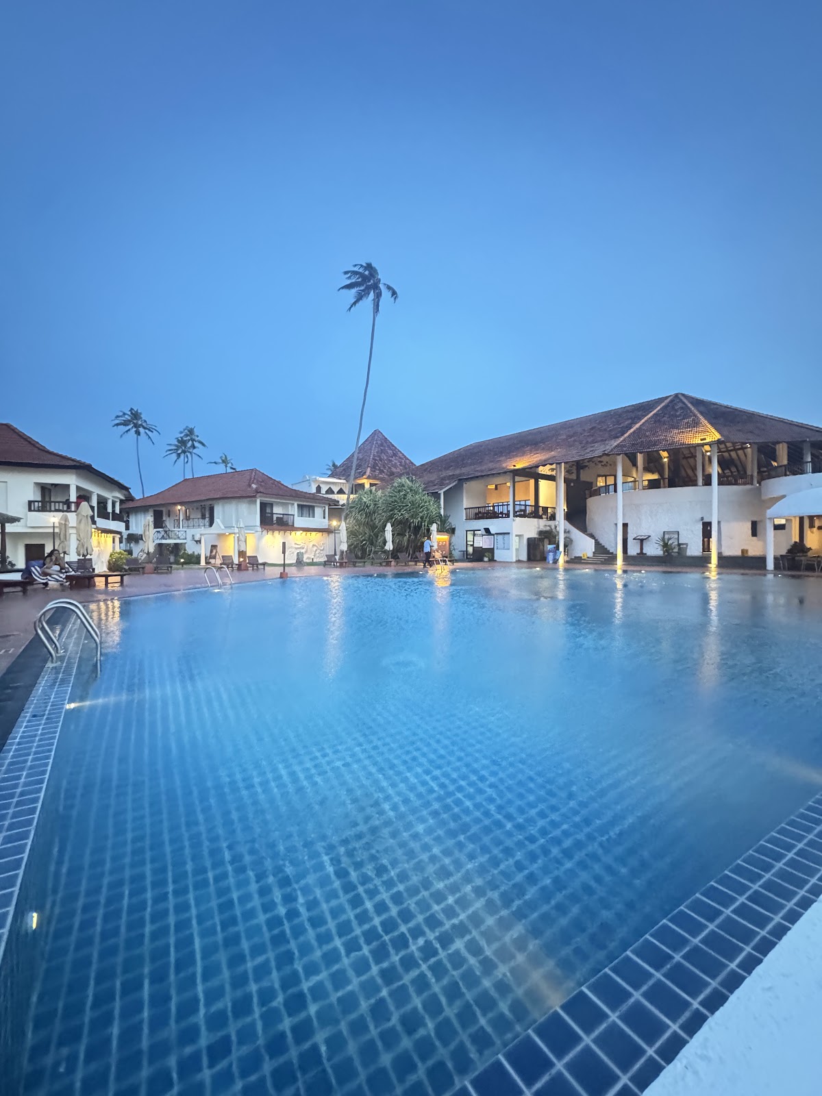 Large swimming pool at dusk with white resort buildings, palm trees, and clear blue sky at Dickwella Resort and Spa.
