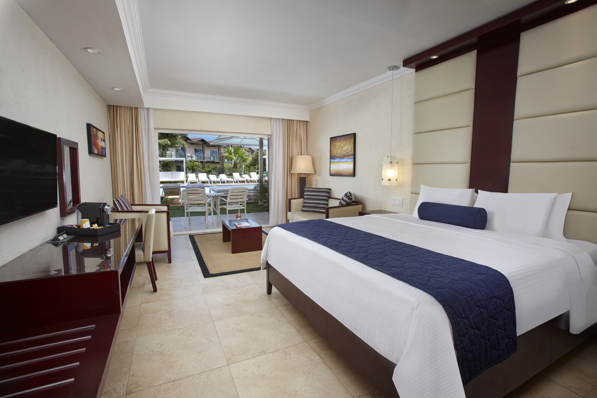 Resort bedroom with king bed, cream headboard, dark navy runner, opening to patio with lounge chairs and ocean view beyond.