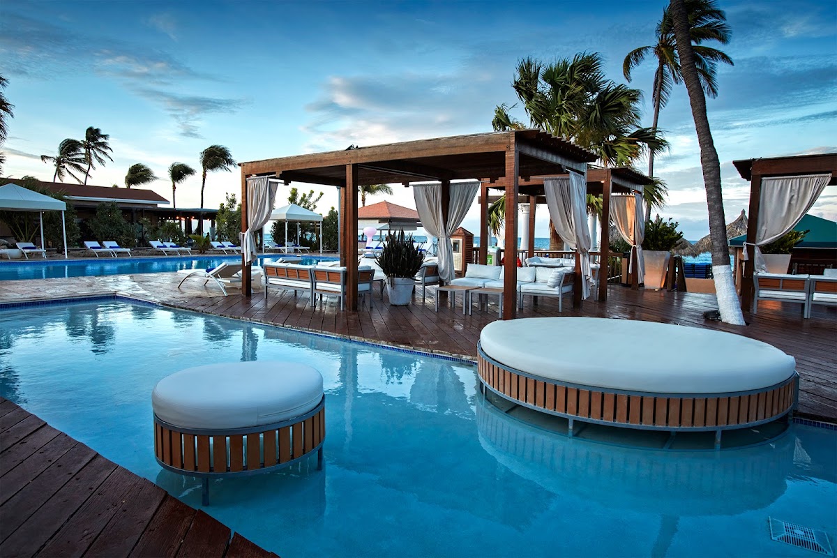 Resort pool with floating loungers and wood pergolas with white curtains, palm trees and ocean visible beyond.