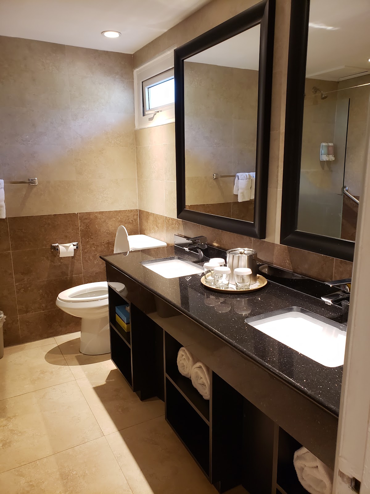 Bathroom with double vanity, black-framed mirror, dark countertop, brown tile, toilet, and amenity tray at Divi Aruba All Inclusive Resort.