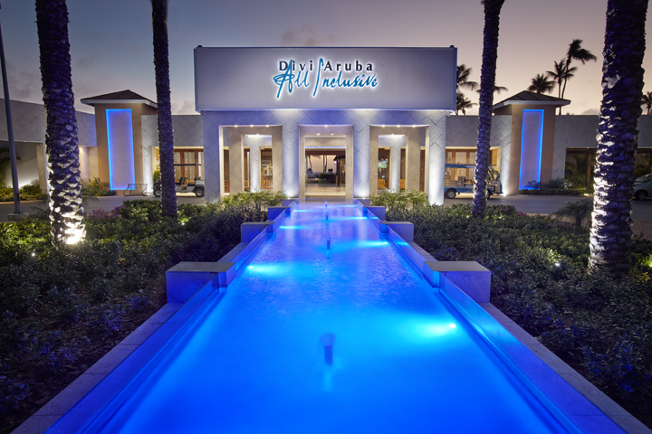 Illuminated blue reflecting pool lined with palm trees leading to the Divi Aruba All Inclusive main entrance at dusk.
