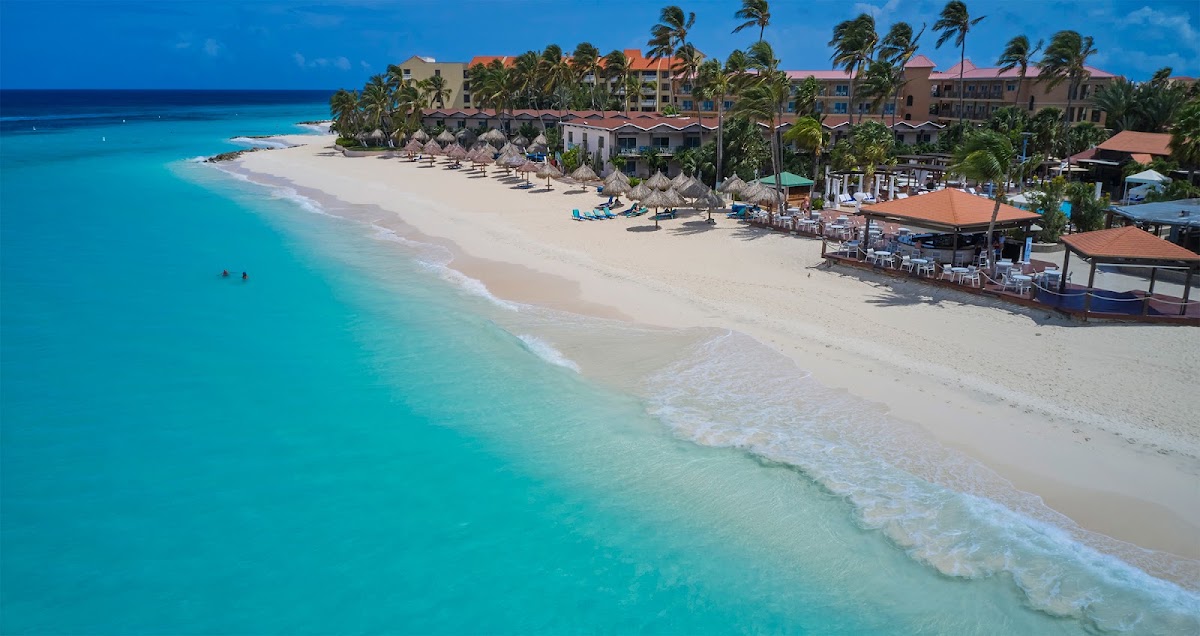 Turquoise waters and white sand beach with rows of umbrellas, lounge chairs, beachfront bar, palm trees, and multi-story resort buildings under clear blue sky.