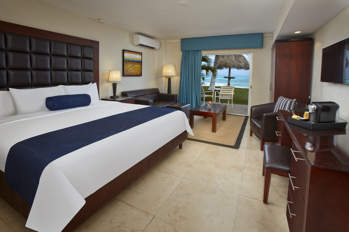 Resort guest room with king bed, sitting area, and ocean view through glass doors opening to beachfront patio with thatched palapa.