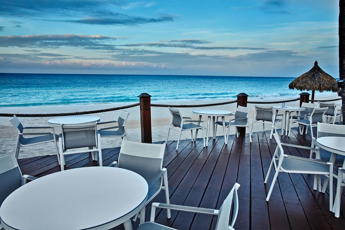 White tables and chairs on a wooden deck overlooking a turquoise beach and ocean at Divi Aruba All Inclusive Resort.