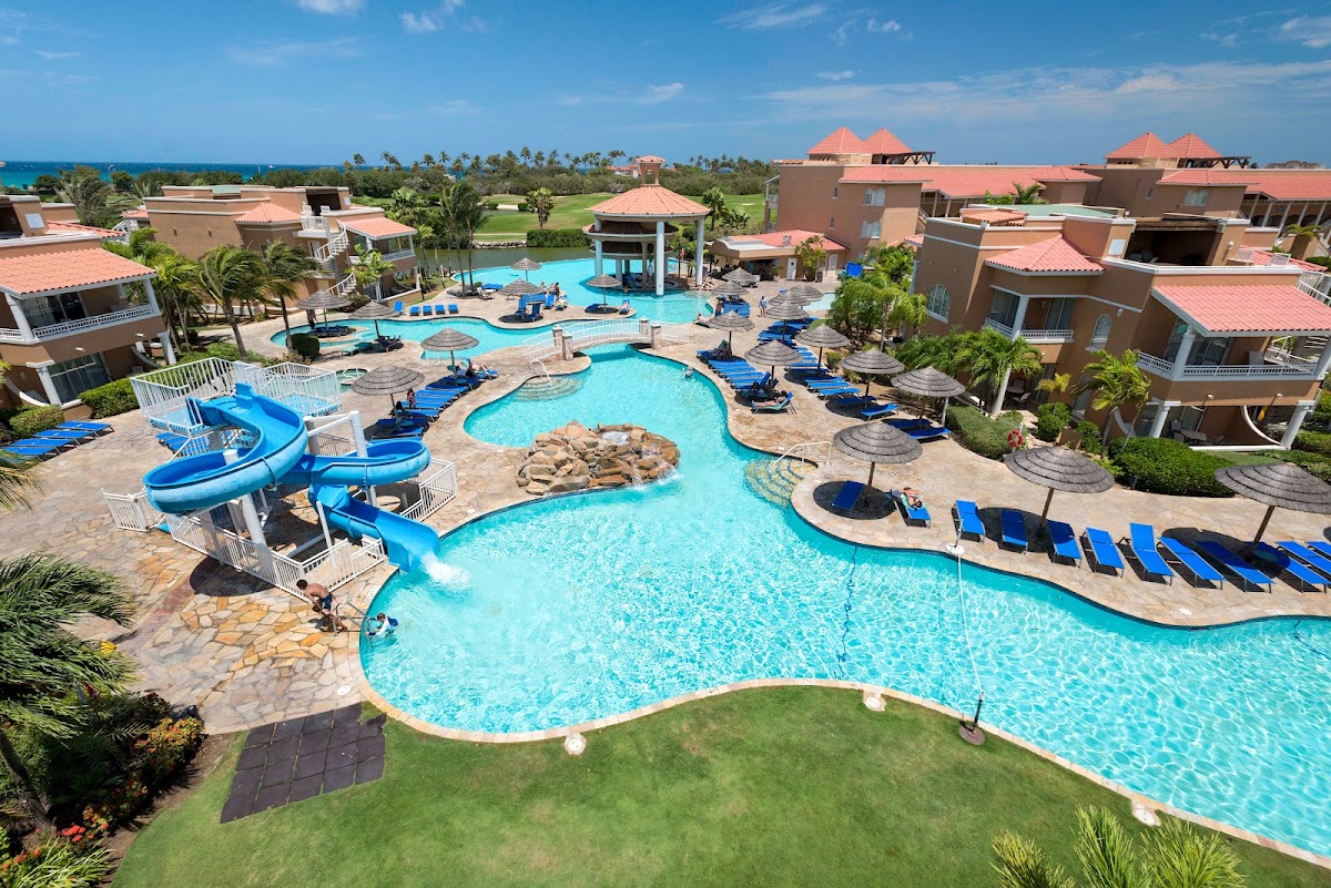 Aerial view of Divi Village Golf & Beach Resort showing curved pools with waterslide, blue loungers, terra cotta buildings with red roofs, and ocean in distance.