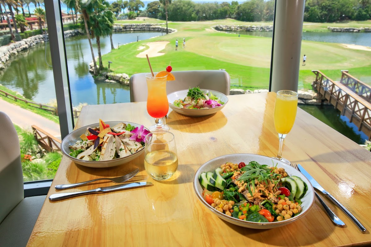 Plated salads and cocktails on a wooden table overlook a golf course and waterway at Divi Village Golf & Beach Resort.