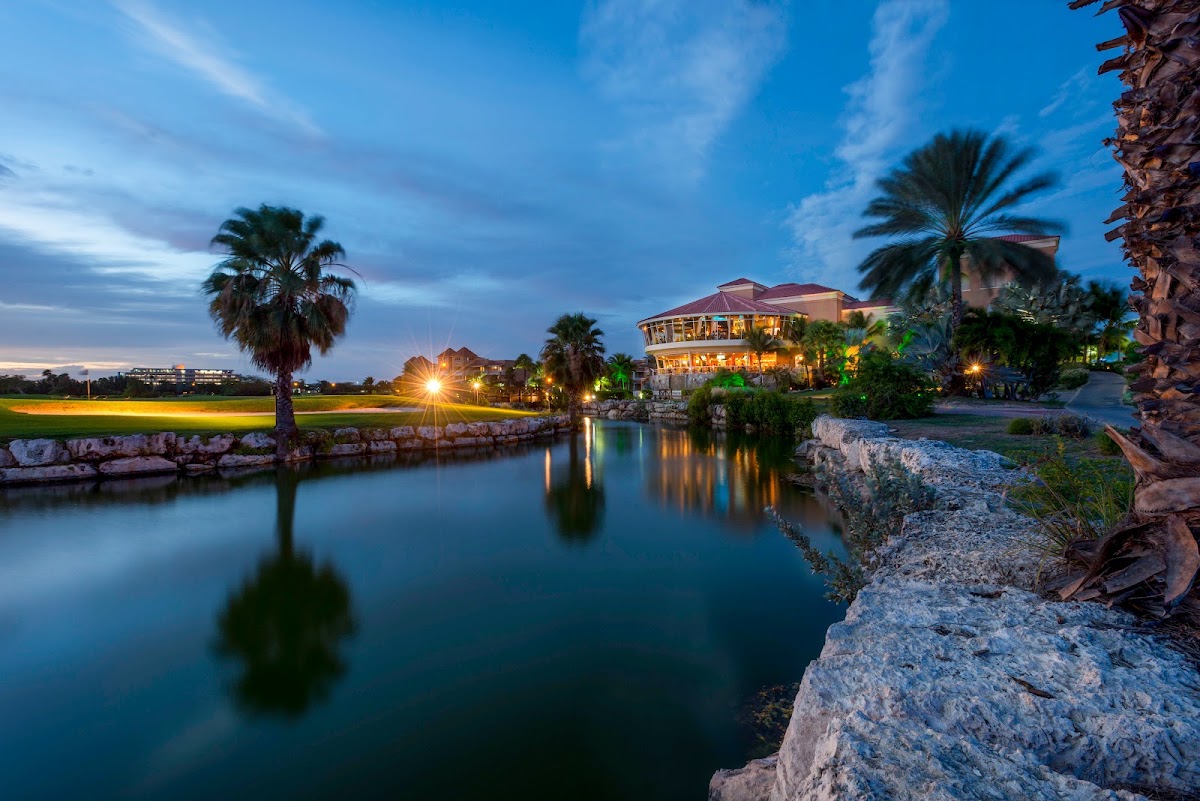 Illuminated resort buildings with palm trees reflected in a calm lagoon at dusk, rocky shoreline in foreground.